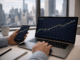 An investor checks rising cryptocurrency charts on a laptop and smartphone with a city skyline visible through the office window.